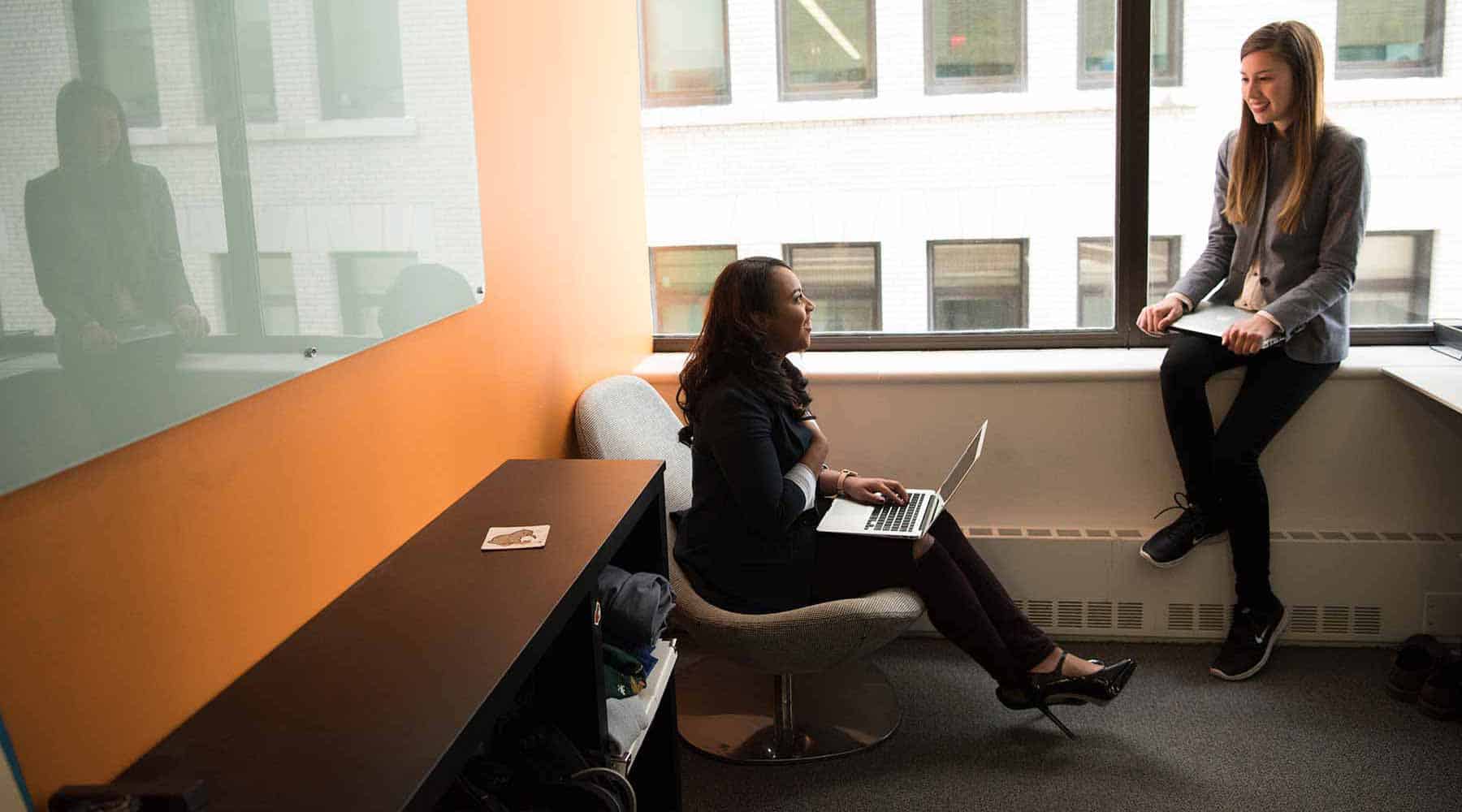 Engaging discussion between two women in an office setting with large windows and modern furniture.